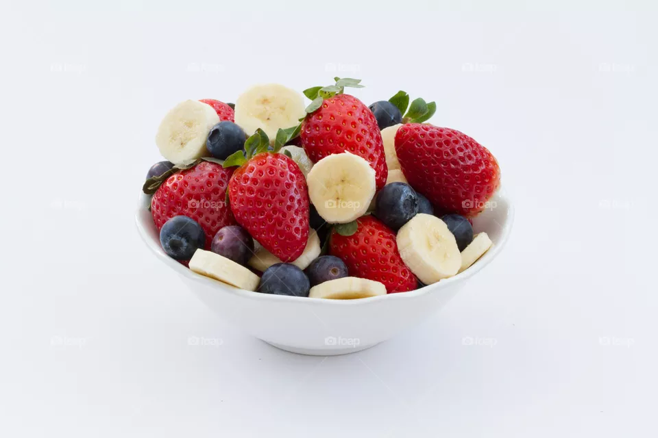 A bowl of fruit including strawberries, bananas and blueberries which are smoothie ingredients on a white background.