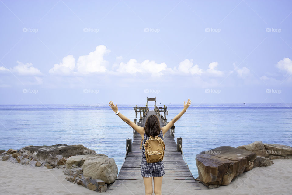 Women raise their arms and shoulder backpack on wooden bridge pier boat in the sea and the bright sky at Koh Kood, Trat in Thailand.