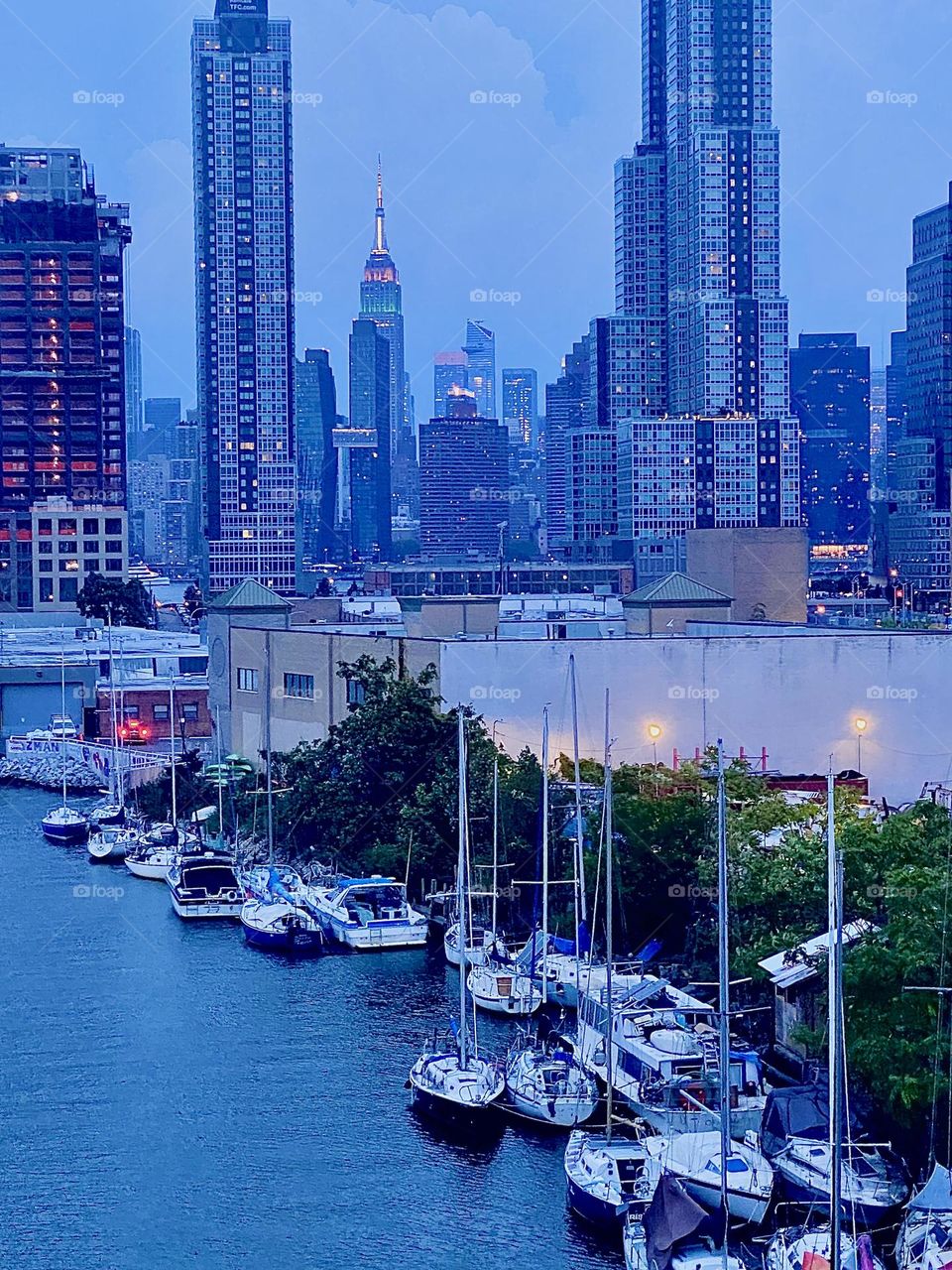 This glorious panoramic view of „Newtown Creek“ at twilight time was photographed from the „Pulaski Bridge“ in LIC, Queens. Behind the nearby skyscrapers we can see „Manhattan“ and the illuminated „Empire State Building“. 2023. Hypnotic Productions