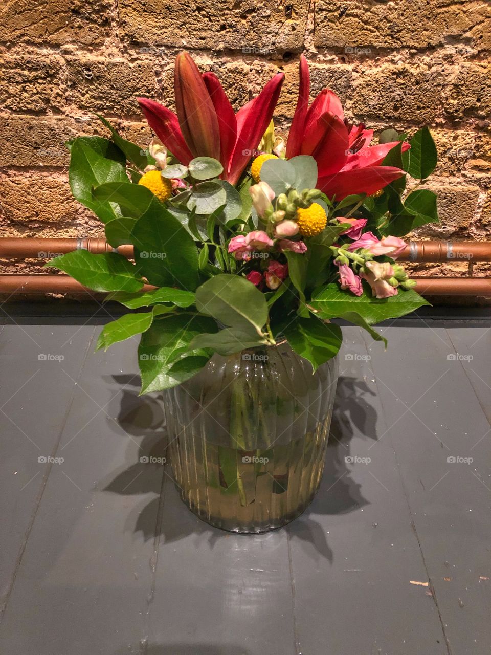 Floral bouquet with red lilies and pink and yellow on painted floorboards in front of exposed brick and copper pipes