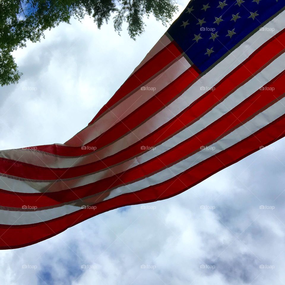 American Flag 🇺🇸 waving in the breeze, looking up at clouds & blue sky. Tree in background if photo.