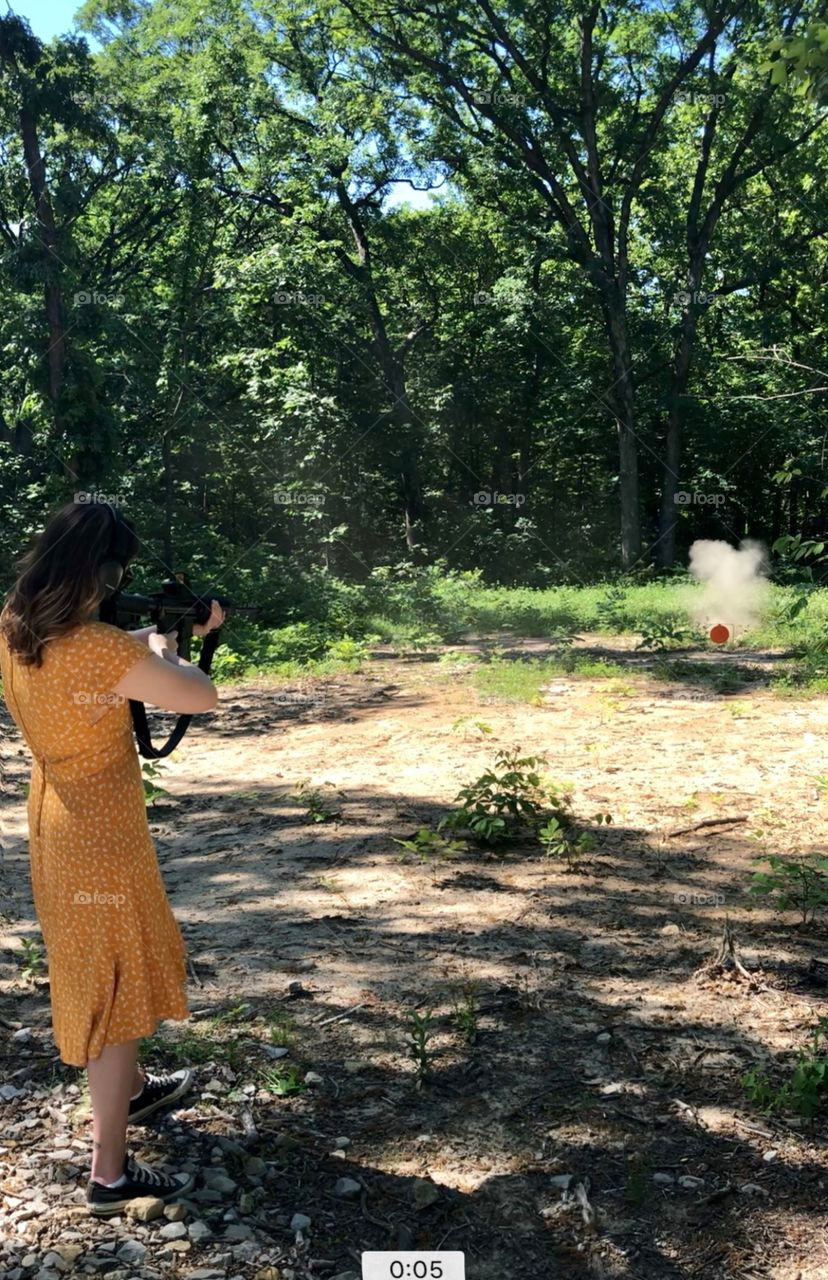 A girl in a yellow dress shooting a large gun in rural Missouri 