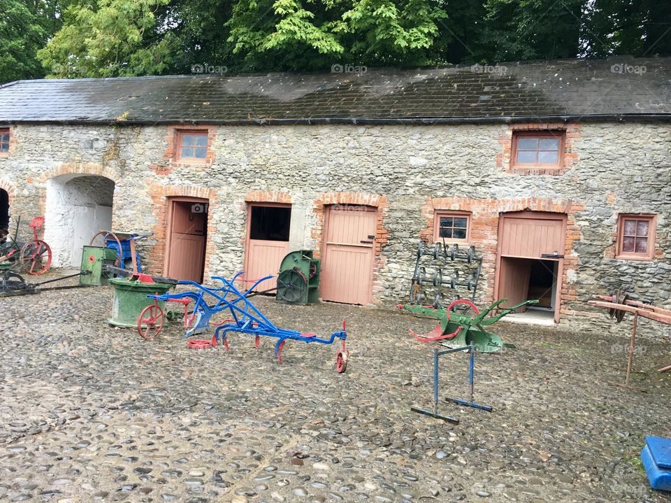 The scene depicts a rustic rural landscape, where vintage farm equipment—painted in vibrant shades of red, blue, and green—stands proudly in front of old Irish barns. The barns are distinguished by windows and doors painted in a soft pink-peach hue.