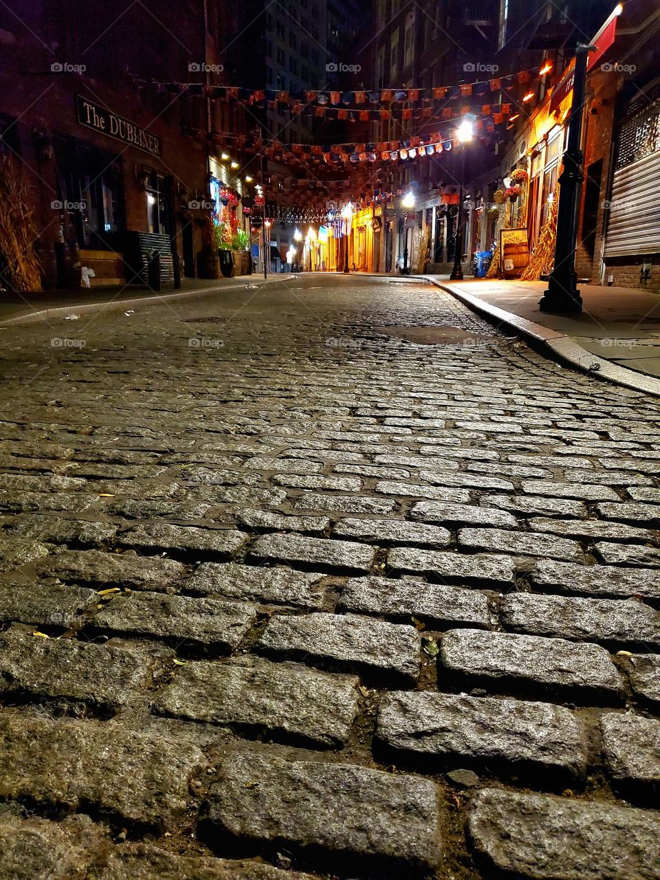 A deserted cobblestone street in New York City glows with eerie light and shadows following a night of fun and revelry
