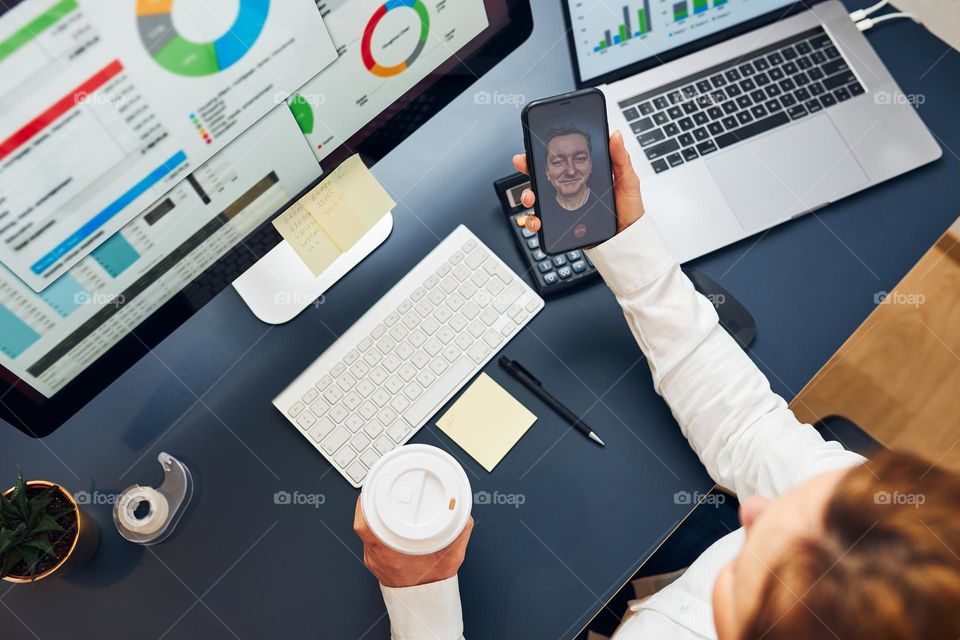 Woman entrepreneur having business conversation, video chat on mobile phone. Businesswoman working with data, charts and graphs on computer screen. Woman holding smartphone sitting at desk in office