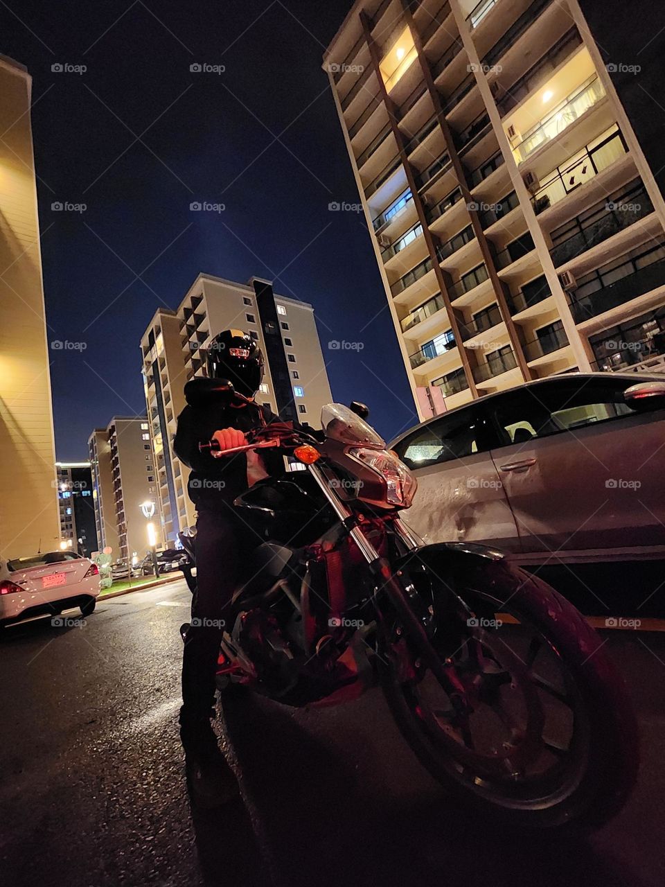 Under the city lights, a rider and their machine stand united—a sleek motorcycle poised for the night’s adventures, framed by towering buildings and the wet shimmer of urban streets.