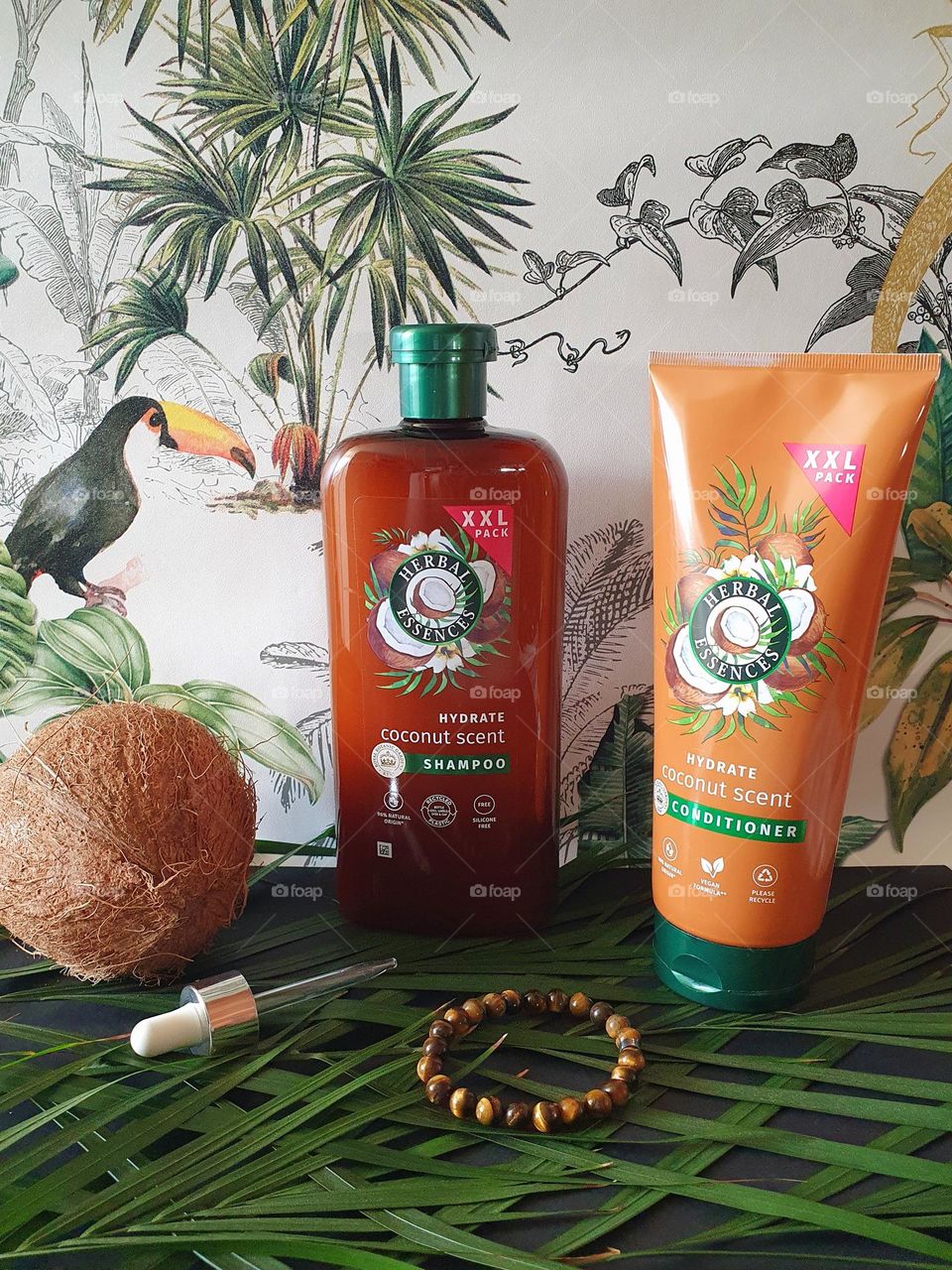 Photo of XXL Herbal Essences Coconut Scent shampoo and conditioner placed on black paper and palm leaves with a tropical paper background together with a coconut, dropper and semi-precious brown beads bracelet