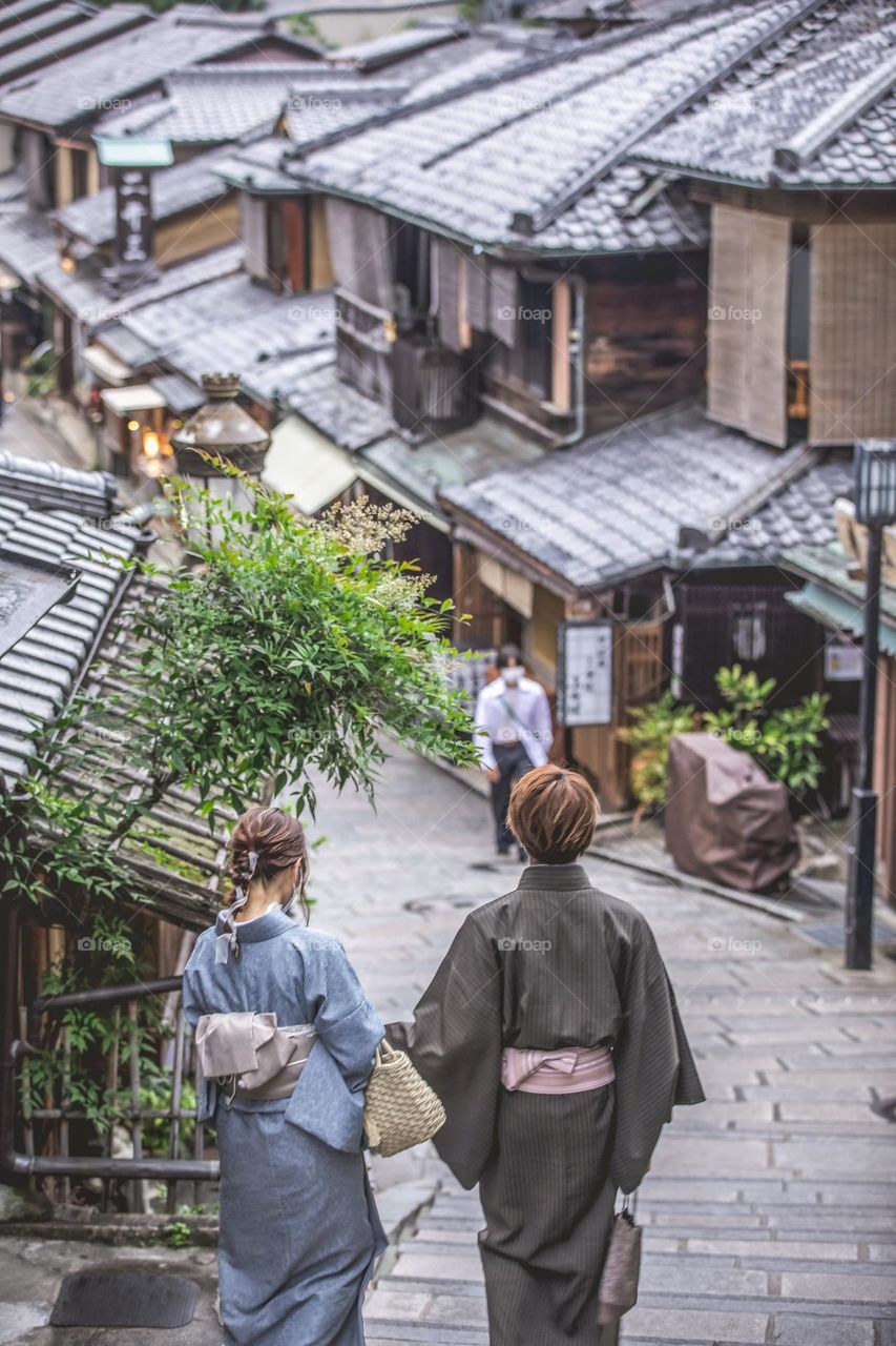 Holding hands, a young couple — a man and a woman — walks through Ninenzaka and Sannenzaka, a historic district of Kyoto, Japan. Both wearing yukata, a summer style kimono. Traditional Japanese clothing. Captured from behind. Scenery in bokeh. 