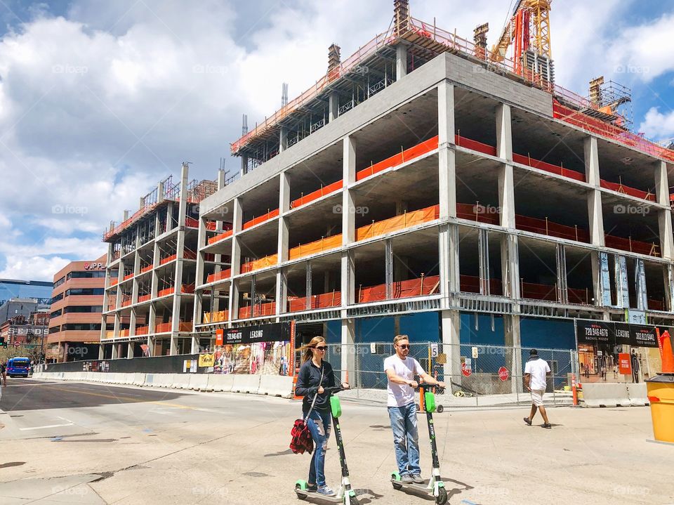 People on electric scooters crossing the road in the city of Denver. There is a building construction at the background