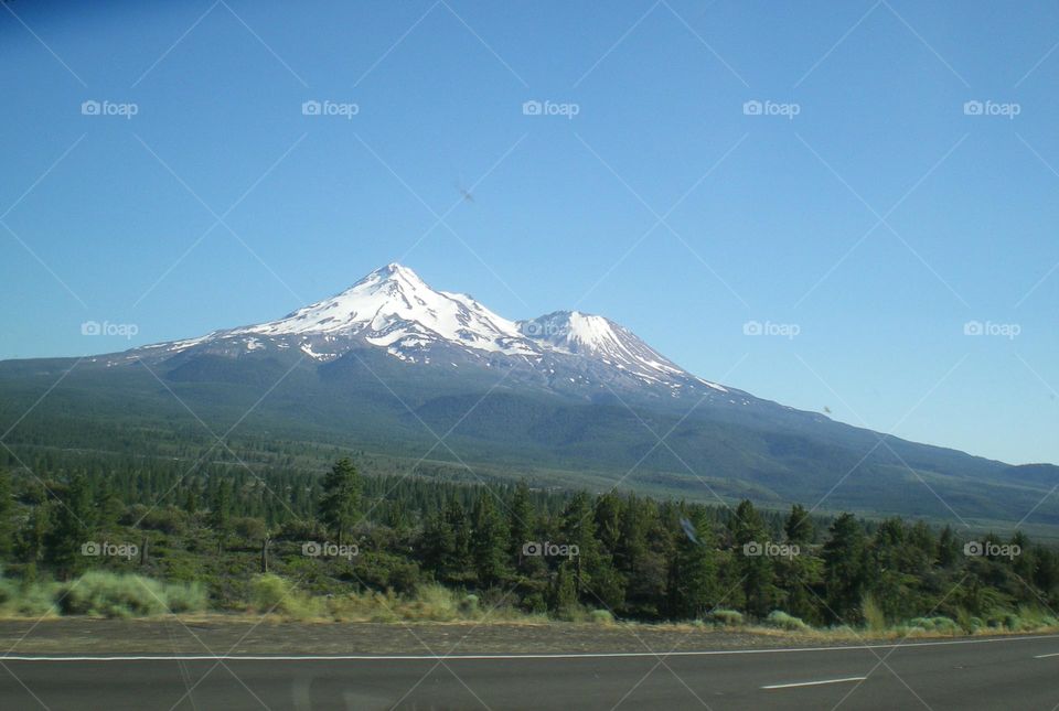 view from a moving car of snow-capped mountains and green trees against clear blue sky on a road trip through California