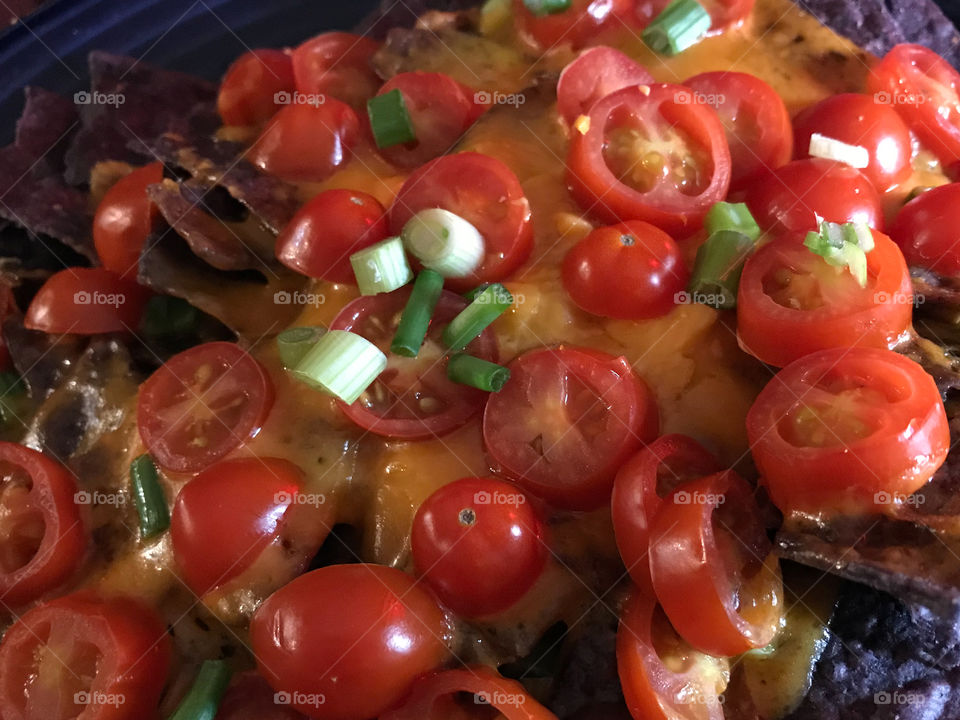 Who doesn’t like Nachos?! Closeup of blue corn chips, smothered with melted cheese, sliced grape tomatoes, green onions, & a splash of hot sauce! Simple pero delicioso!