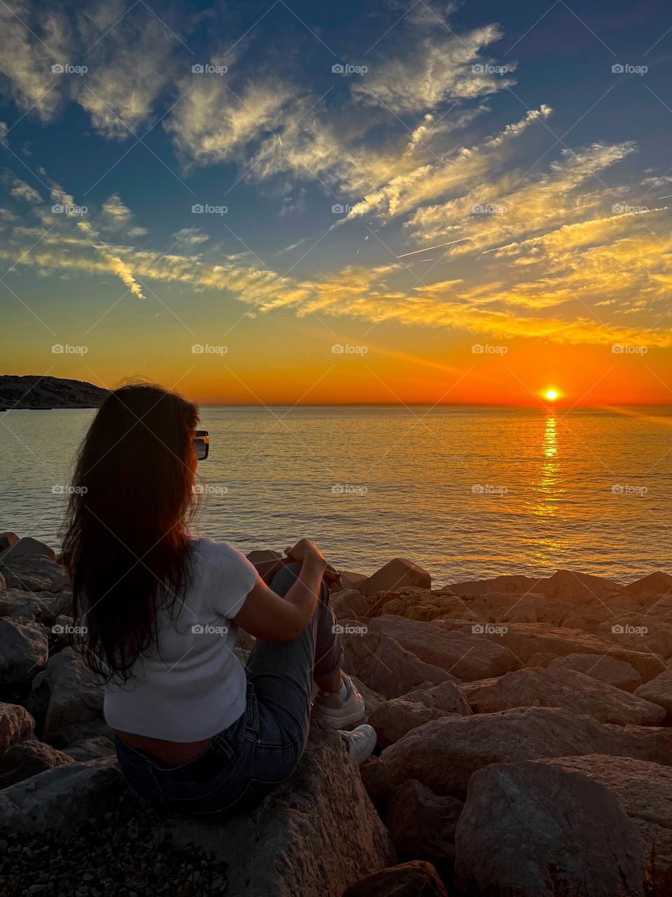 Portrait of a beautiful young caucasian brunette girl in jeans, a belry t-shirt and sneakers sits from the back on the coastal boulders near the mediterranean sea and admires the unusual spectacular sunset on the horizon on the island of frioul in fr