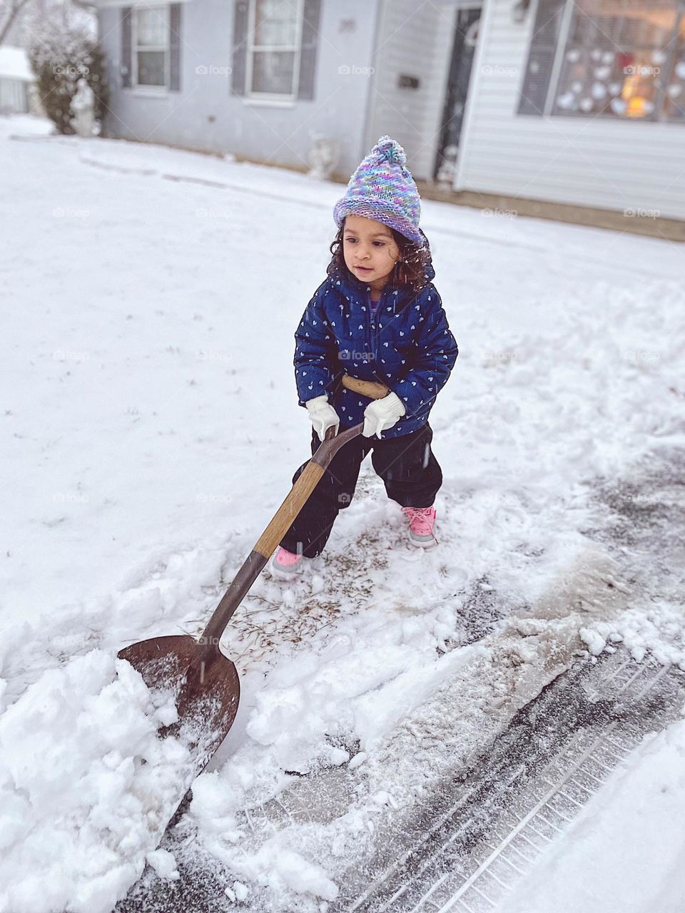 Toddler shoveling snow in the driveway, toddler helps with chores, toddler clearing piles of snow, cleaning up after a winter storm, toddler dressed in warm winter clothes, snow shoveling in the winter, winter wonderland 