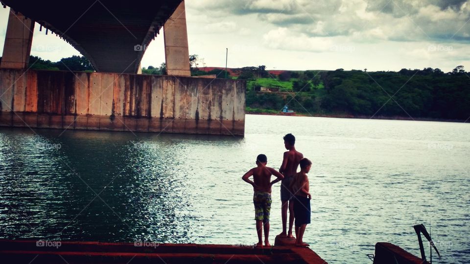 Crianças sob uma ponte de concreto, à beira das aguas do rio.
