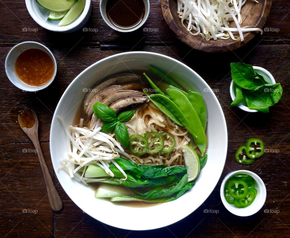 Vegetarian Pho Soup with snow peas, boo choy and mushrooms in a white bowl on a wood table with ingredients.