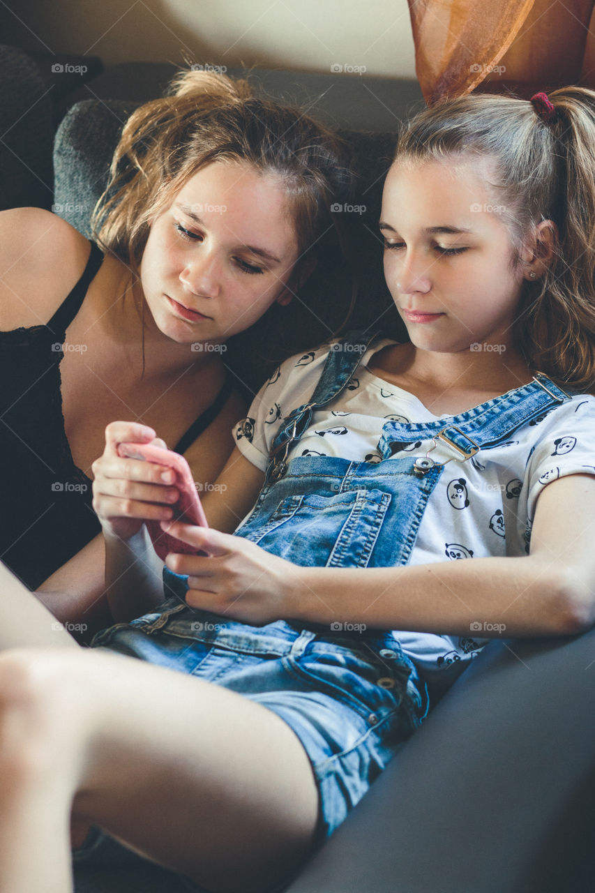 Young women using mobile phones watching music clip, texting, messaging. Teenagers using the smartphones, sitting on sofa at home. Using technology devices. Girls wearing summer clothing