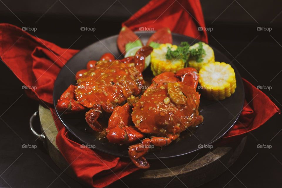 Crab flavored in creamy garlic sauce served with corn, tomatoes, lemon wedges and cherries on a black plate set on a red cloth set in a wooden setting. Asian flavor crab.