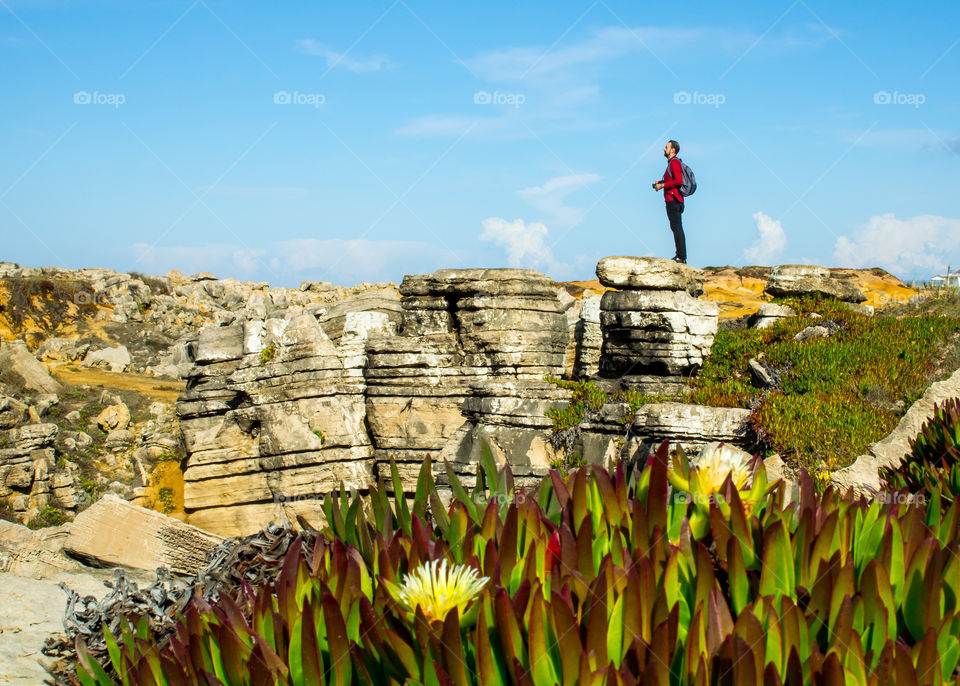A man stands upon cliffs looking out and up to the blue sky surrounding him.