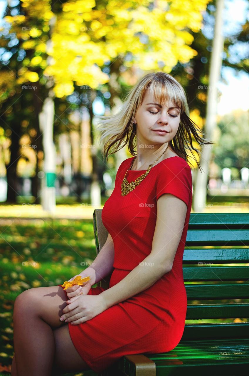 Portrait of beautiful young blonde woman, girl in red dress sitting at the bench in the autumn park