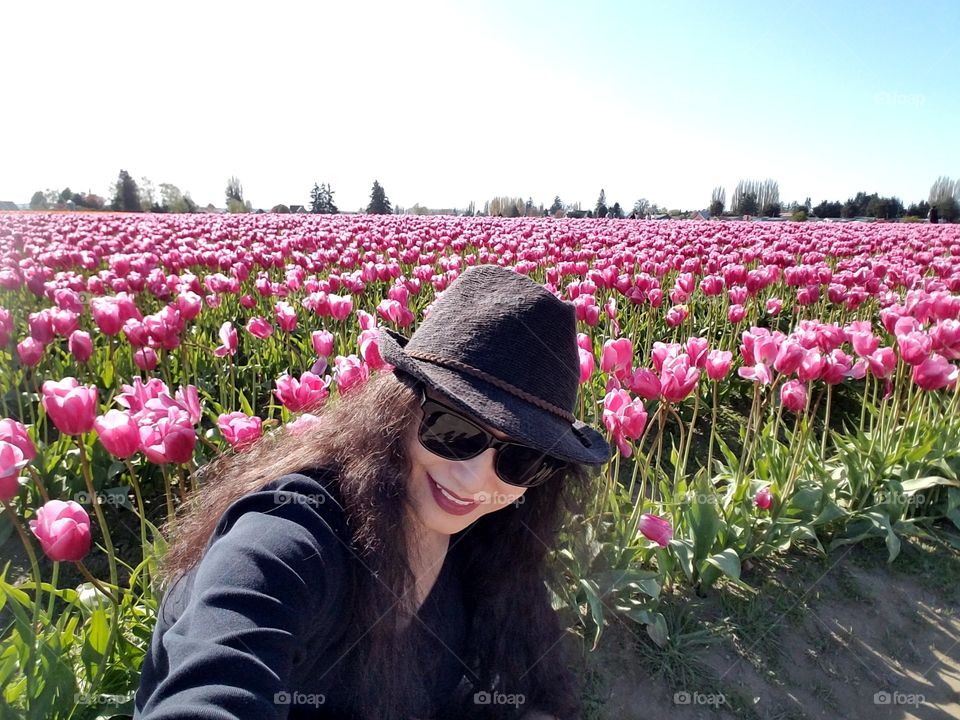 A long black hair selfie wearing sunglasses with black cowboy hat and black shirt in a pink tulip field.