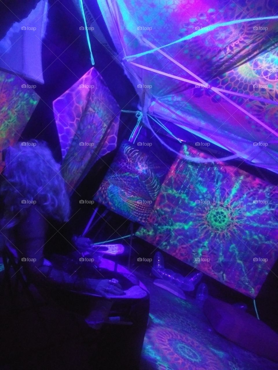 Future Witch Teleportation. At a Halloween party, a lone witch relaxes and gazes through the psychedelic scene of art around her, brought to a different kind of life under a blacklight. She is going places in her mind.