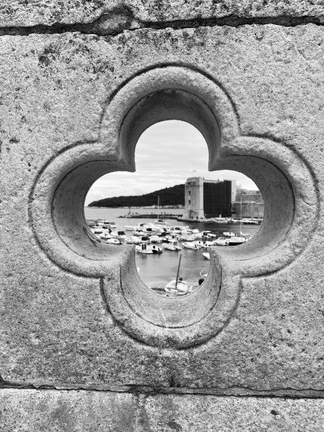 Black and white photo looking through the stone fence at the view of Dubrovnik’s old harbor in Old Town.