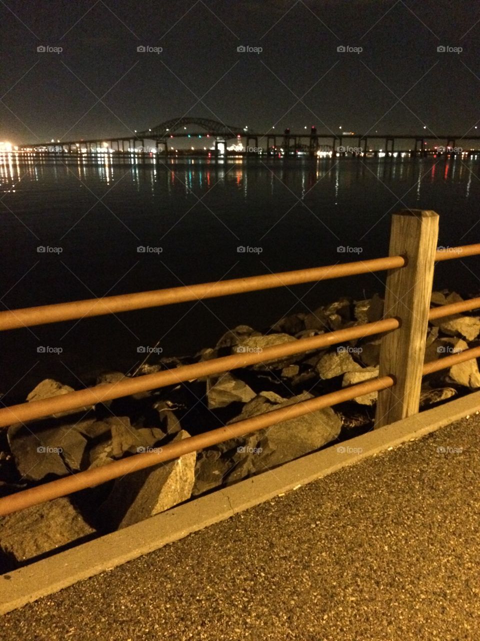 Fenced path with lights of nighttime city and bridge in background across water