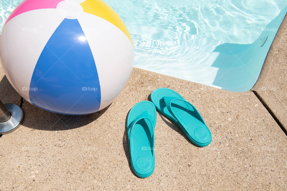 Close-up of a beach ball and pair of women's flip flop sandals next to a swimming pool