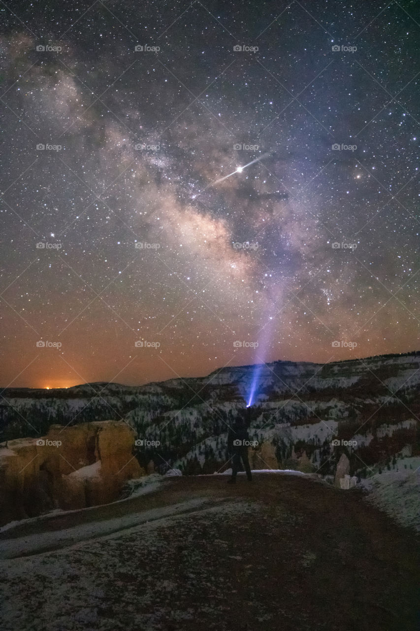 A lone hiker with a flashlight pointing up into the night sky, with the Milky Way brightly visible over Bryce Canyon National Park. 