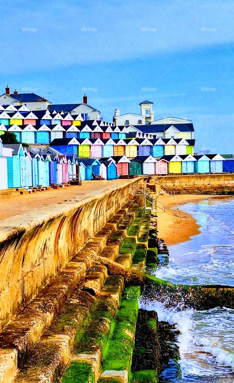 Sunlit tiers of bright coloured beach huts with a blue sky and surf breaking on a honey coloured promenade with bright green seaweed