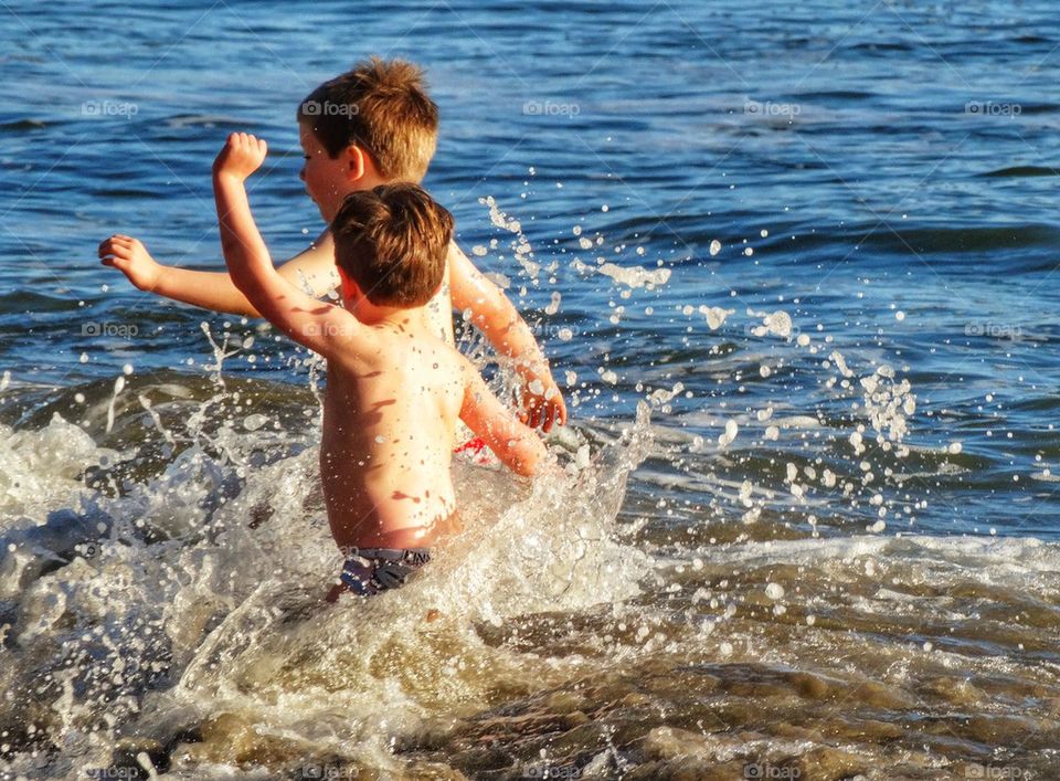 Two Young Boys Challenge The Ocean. Boys Splashing In The Ocean

