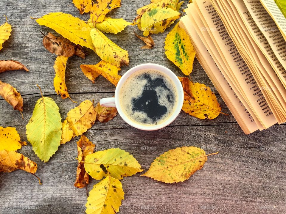 Cup of coffee on wooden table with a book and yellow autumn leaves beside