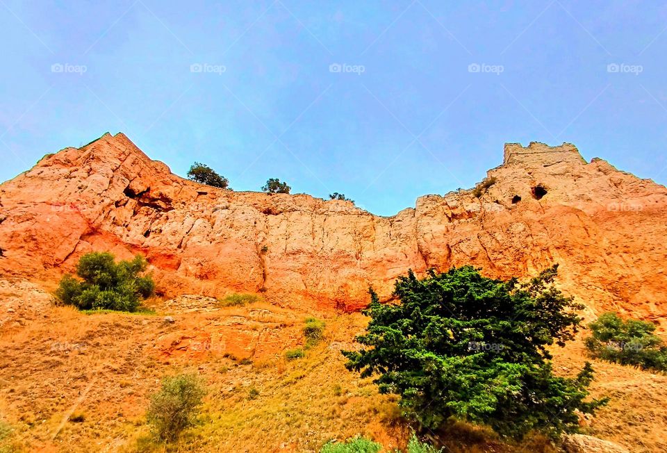 the Roucas of Vitrolles, a striking natural rock formation characterized by its reddish-orange hues. The cliff face rises prominently against a clear blue sky, with rugged textures and crevices that add depth and interest to the scene.