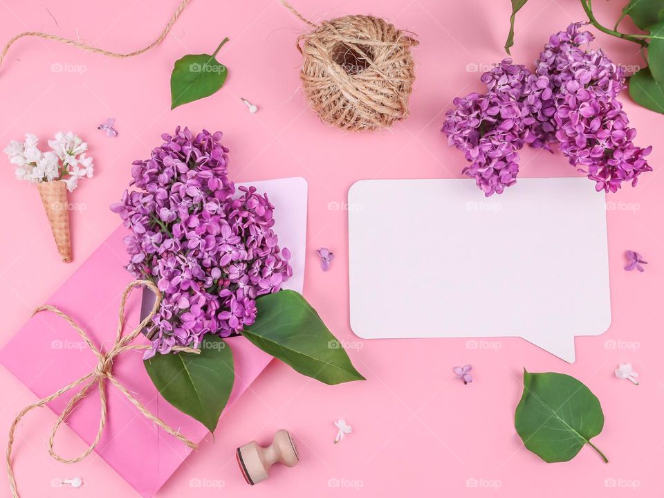 Envelope with lilac branch, empty white callout with wooden seal, jute thread, waffle cone and scattered flowers on a pink background with copy space in the center, flat lay close-up.