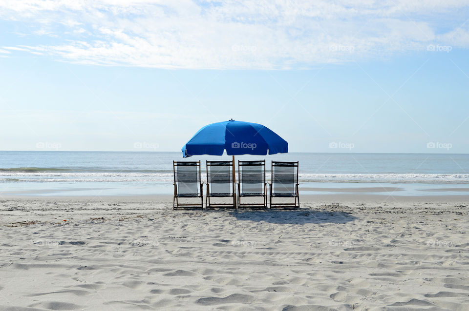 Isolated row of blue lounge chairs and umbrella in the sand of a beach overlooking the ocean and blue sky
