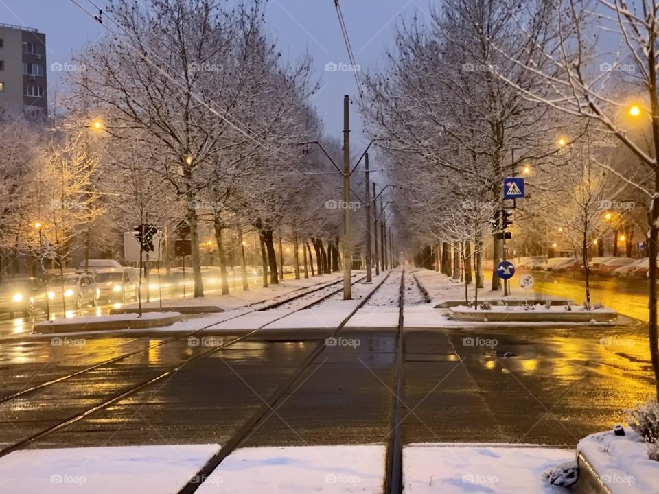 Illuminated streets by cars with headlights on captured in long exposure and in the middle, two tram tracks covered by snow, in the early morning.