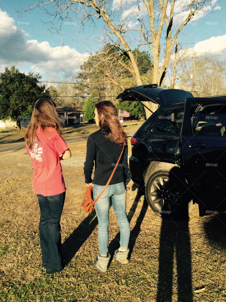 Two young women outdoors standing with their backs to the camera and a vehicle showing.