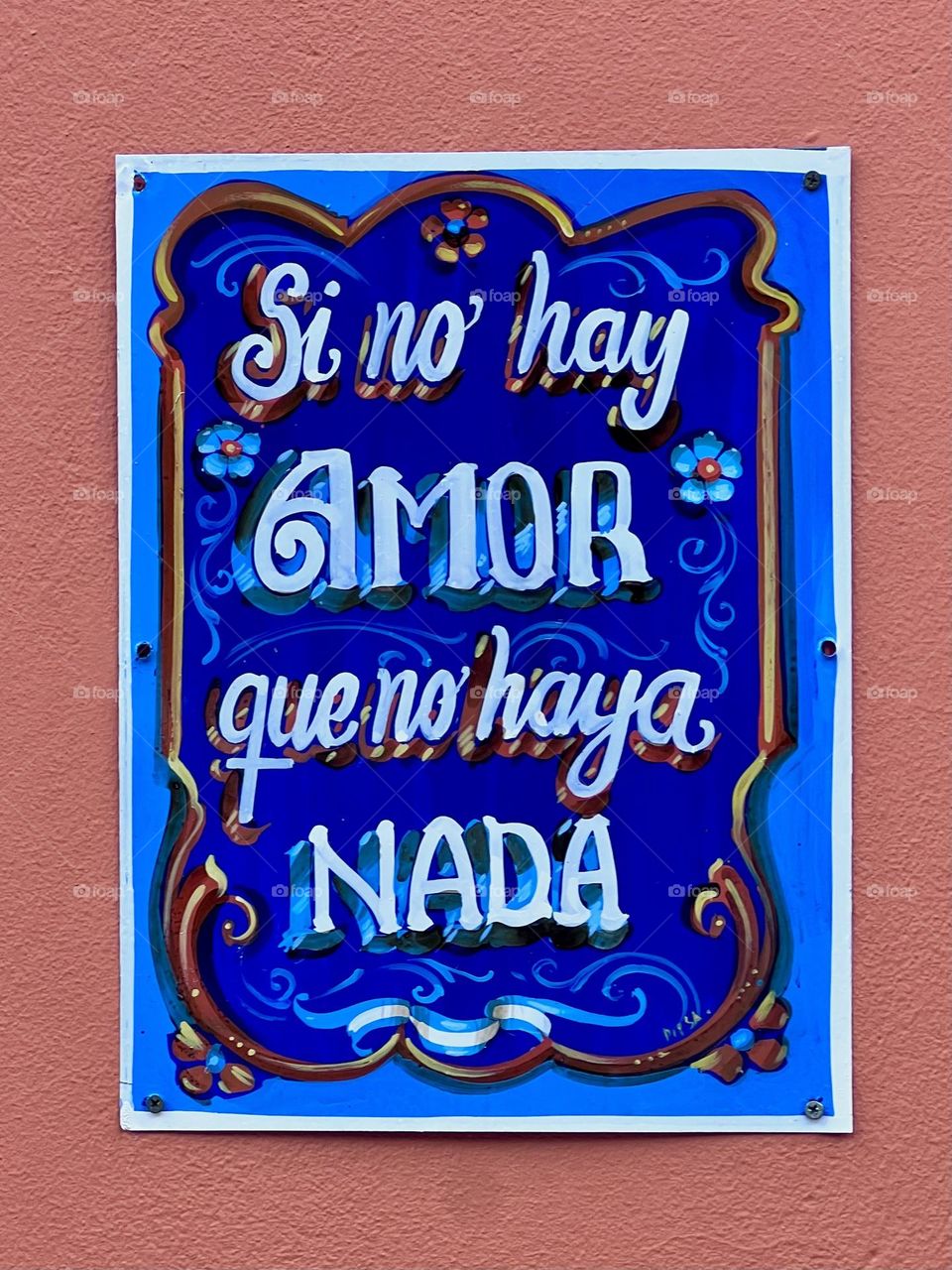 A vibrant and colorful sign with the phrase 'Si no hay amor que no haya nada', expressing the importance of love in our lives. The vibrant blue background contrasts with white letters and floral details, evoking a feeling of joy and hope.