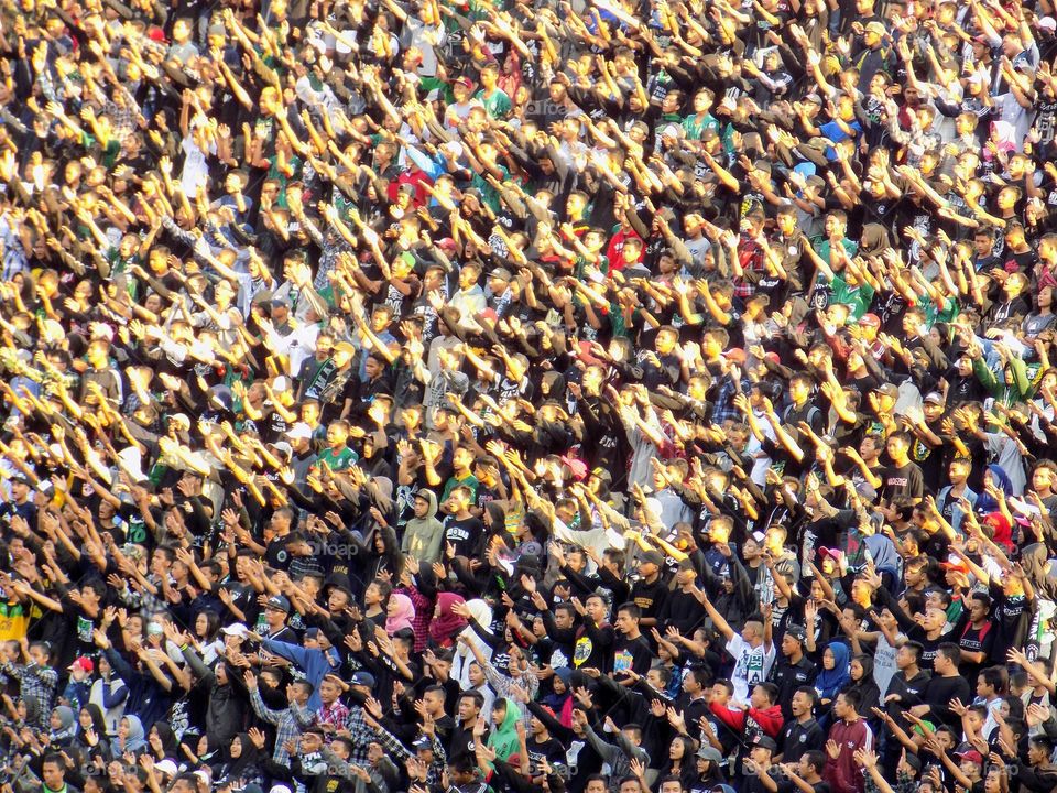 A crowd of people watching a football match in the afternoon at a football stadium, they are raising their hands to shout and support their favorite team to compete