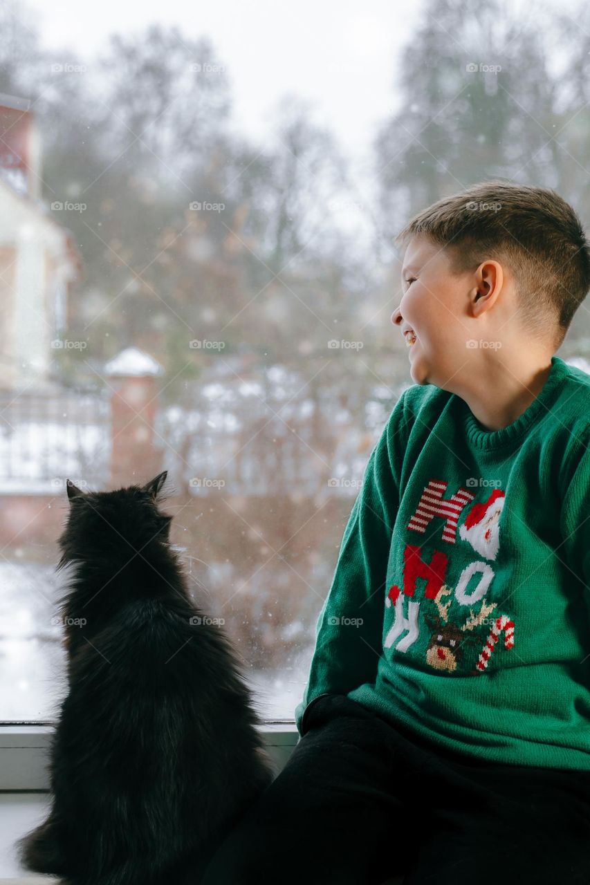 Cute smiling teen boy looking through the window in the winter with black cat 