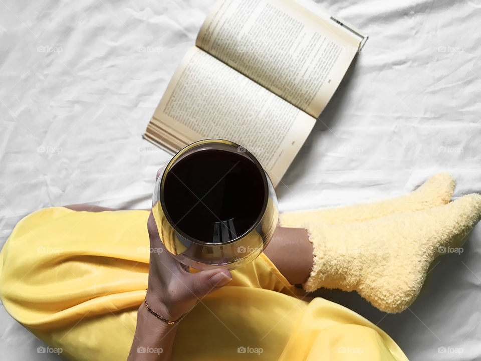 Young woman enjoying wine and reading book in cozy yellow socks in bed in winter 