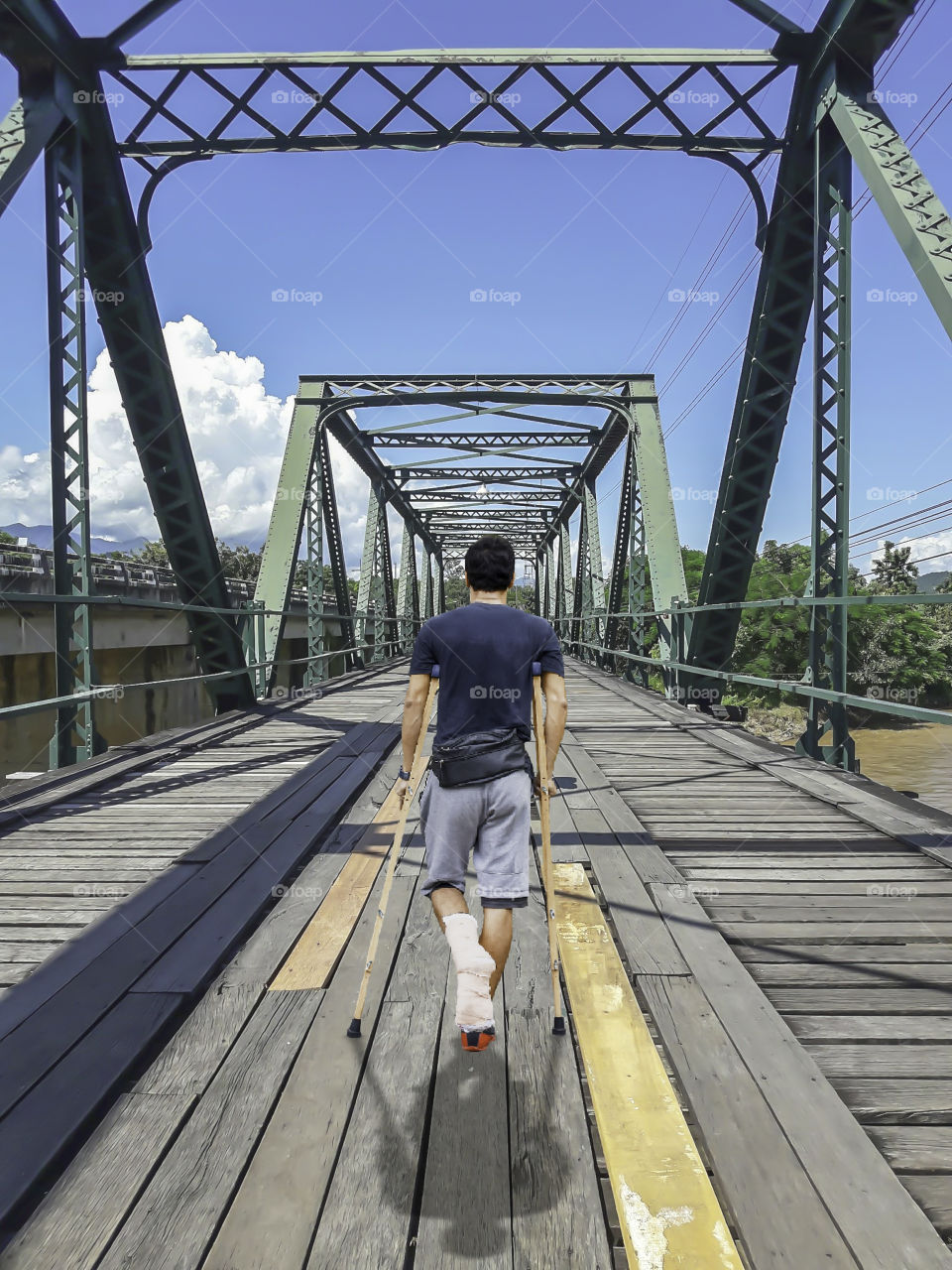 Asian man used crutches walks on The wooden bridge steel structure across the Pai River, Phitsanulok in Thailand.