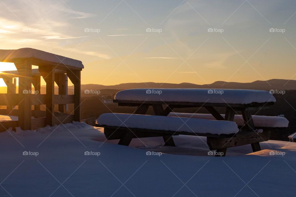 Sunset on a mountain top in the middle of winter in New England. Snow covered railing and picnic table with a beautiful sky.