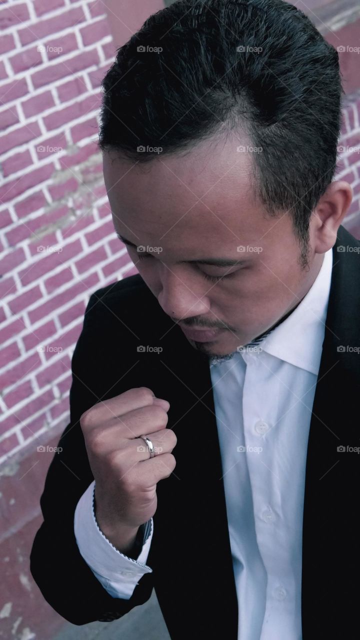 Close up of an Asian man dressed in a black suit with his right hand clenched into a fist with a wedding ring on his ring finger, a flat expression of happiness.