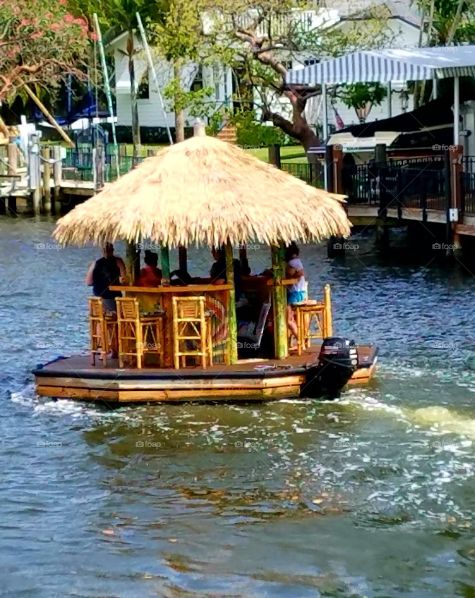 Thatched wooden round Bar Boat with engine going down the river. Bar stoole all around, no cars seen. People drinking safely. Funny pic on ocean through way.