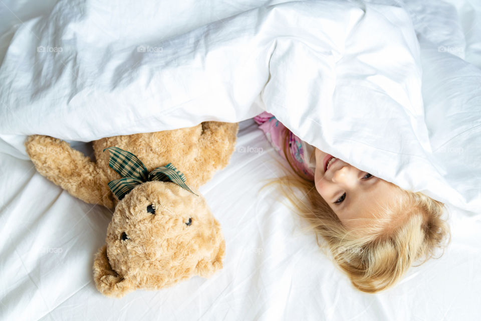 Happy little girl with blonde hair lying on the bed with teddy bear 