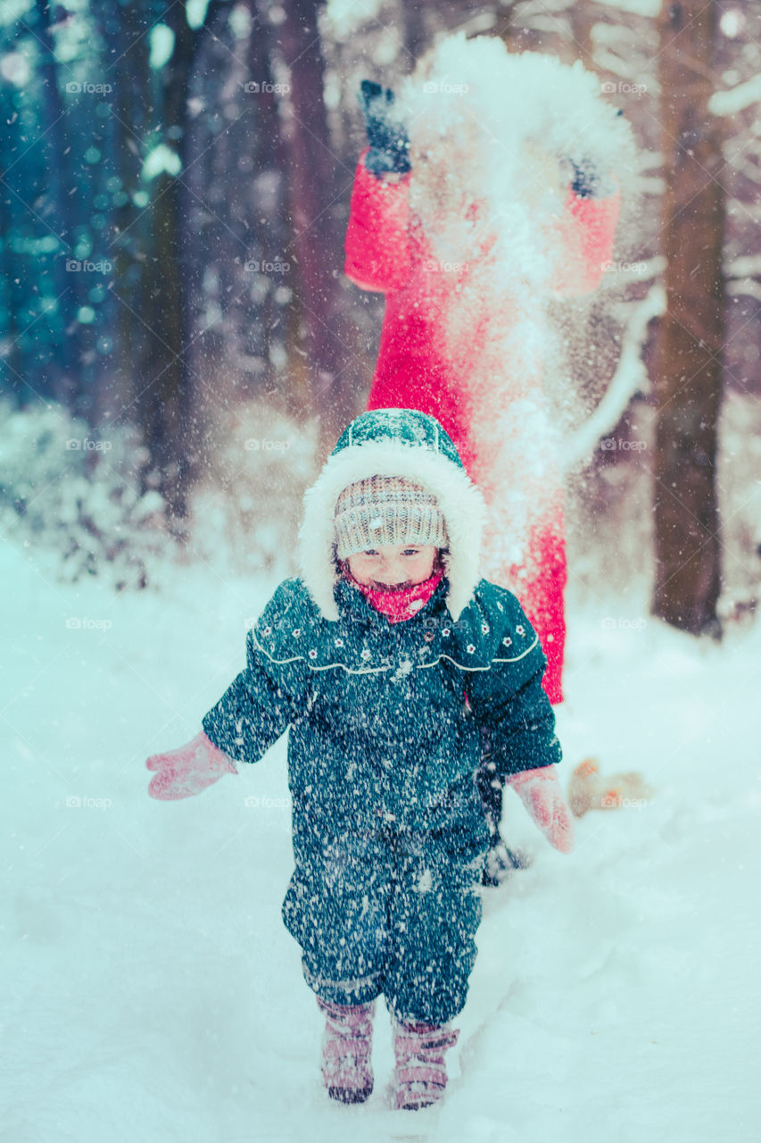 Mother is playing with her little daughter outdoors on wintery day. Woman is throwing snow on her child. Family spending time together enjoying wintertime. Woman is wearing red coat and wool cap, toddler is wearing dark blue snowsuit