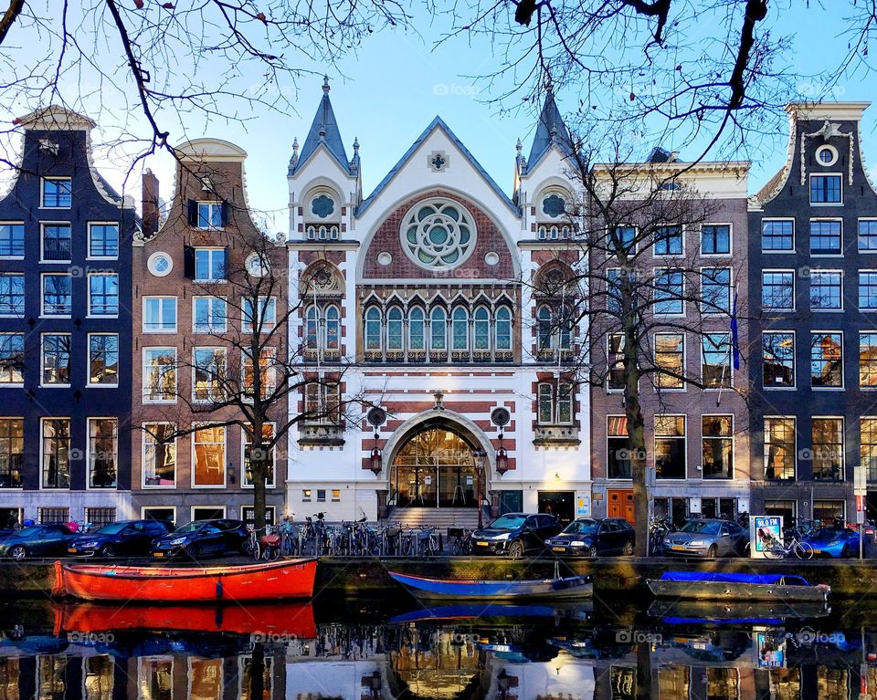 An historic church, with row houses on either side, lines a canal in Amsterdam. Various small boats float on the water in the foreground. 