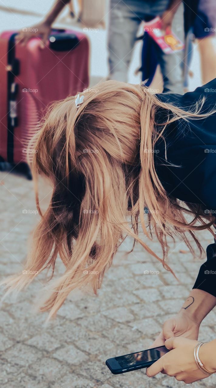 Caucasian blonde girl with long hair stands in a tilt and holds a mobile phone in her hands typing an urgent message with her fingers against the background of a blurred street with a suitcase and people in line, side view close-up.