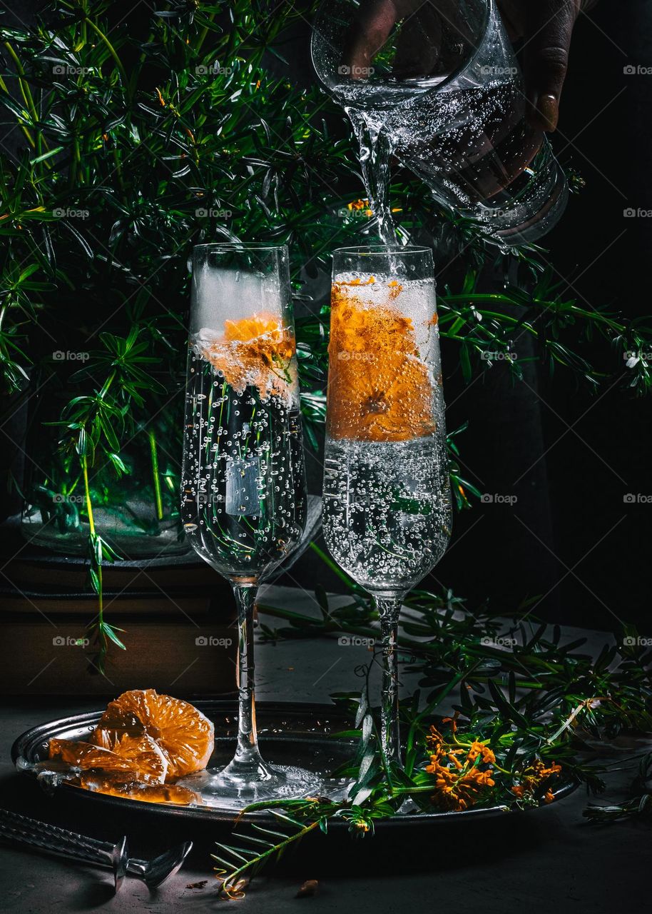 Two glasses of champagne shot in a low key stand on a silver plate surrounded by slices of tangerine and leaves of fragrant herbs - luxury lifestyle